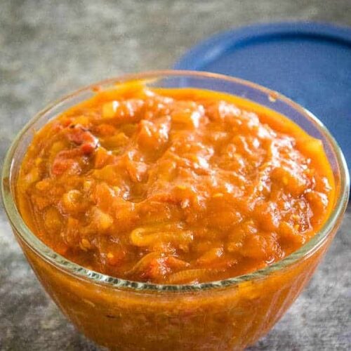 Tomato Chutney Recipe in a clear glass bowl for storage with a blue lid in the background.